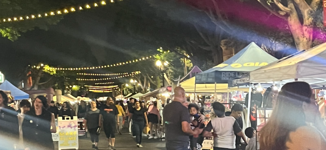 During nighttime, there are a crowd of people walking with some standing and talking to people at various stands. None of the names are readable but the photo has some streaks of pink and blue due to lighting. There are several strings of lights that slowly start fading away in the top right corner.