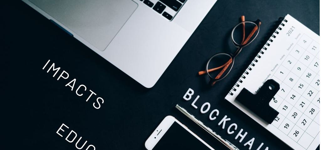 Image of a computer, a note book and a phone on a table with the words : Blockchain, Impacts and Education, in upper case letters written on the table