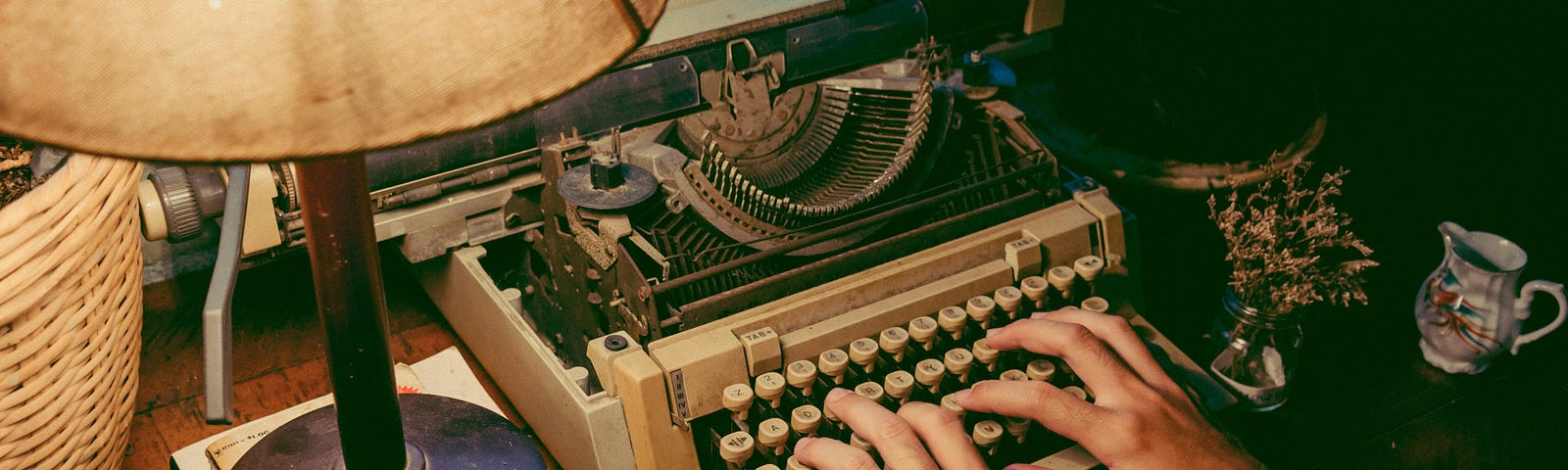 Two hands on an old typewriter, writing at a small, cluttered desk.