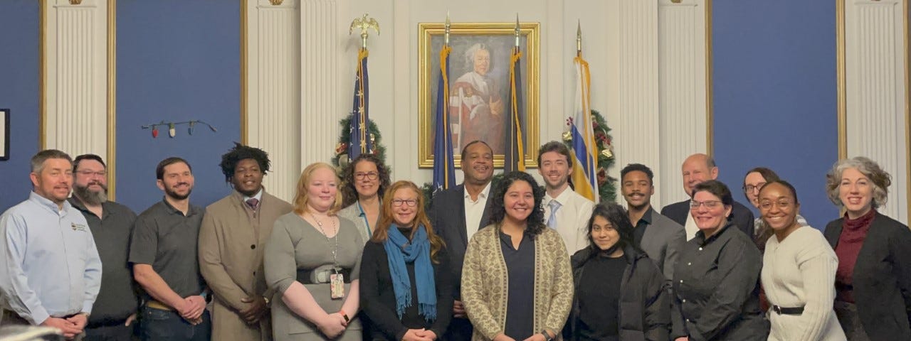 17 members of city government and companies standing, smiling, and posing together in a decorated city conference room