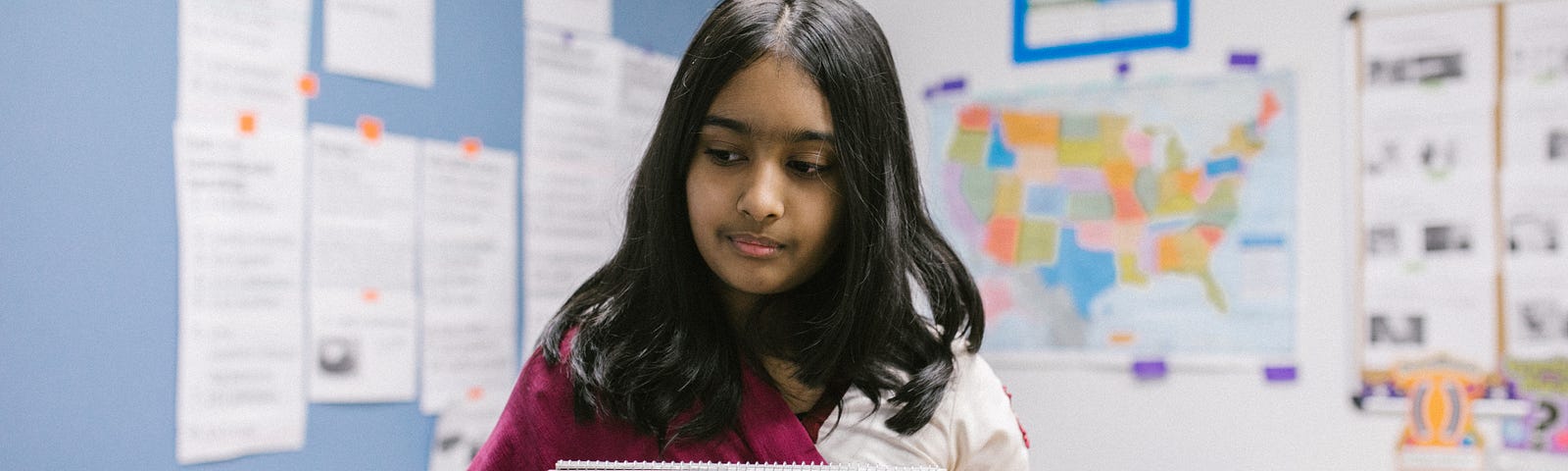 Indian girl in a classroom wearing a pink and white sari, holding up an open notebook with the words “stop the bullying” written across the page in red marker