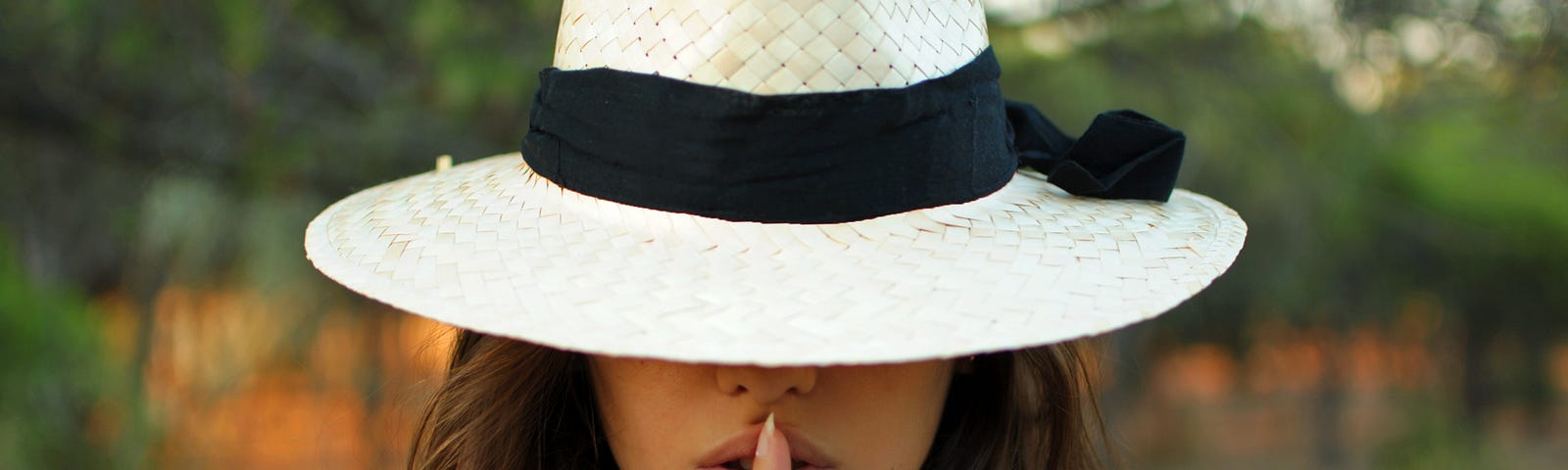 A close up of a white women’s face. A large brimmed hat covers her eyes, and she has a finger on her lips to shush the audience.