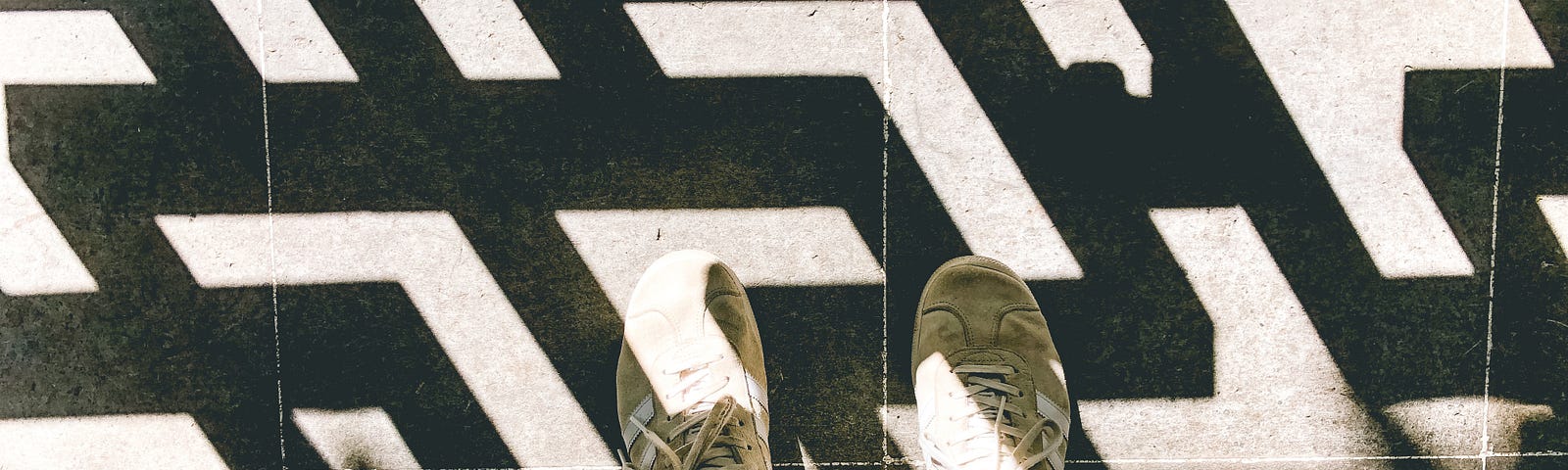 Photo shot down towards the feet. Blue jeans and cream shoes are interrupted by a geometric shadow along the floor.