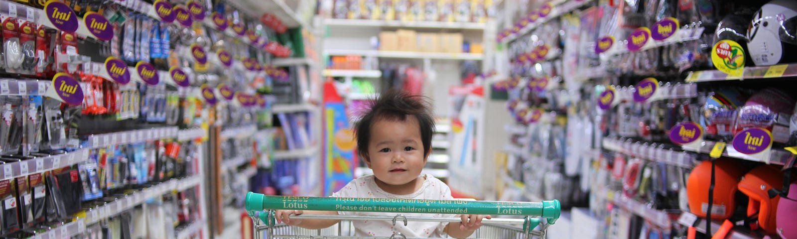 A baby sitting in an empty shopping trolley in an aisle of a department store with shelves full of colourful items.