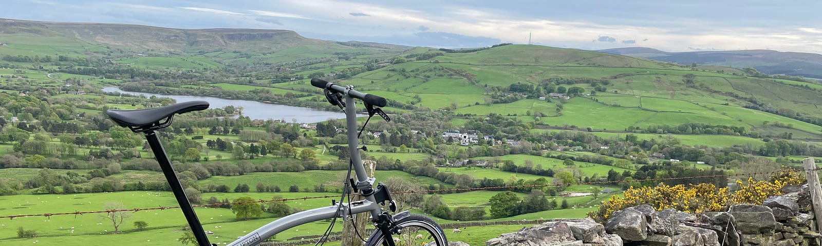 A Brompton bicycle balanced on a stone fence with a green Peak District valley in the background as well as a body of water