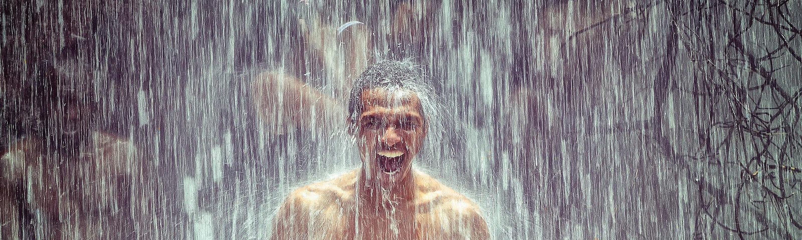 A man with no shirt is up to his waist in water while heavy rain pours over him. He is smiling.