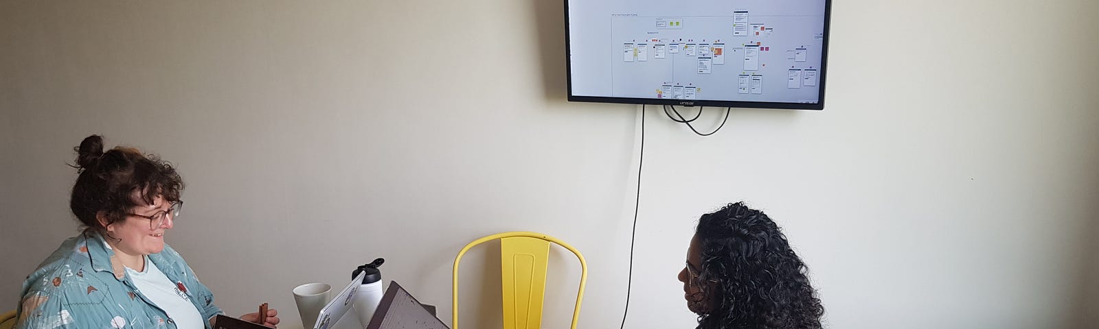 A picture of our work setup. There is a table with 3 laptops on it; 2 people are sat around the table working on their laptops. On the wall there is a tv screen, which is presenting images from one of the laptops.