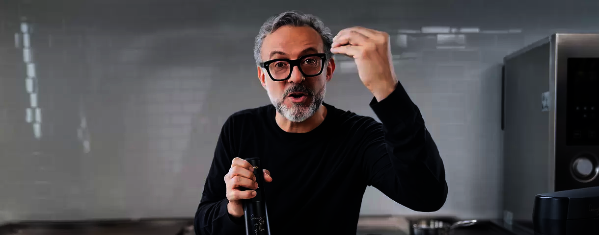 A person with short salt-and-pepper gray hair + matching facial hair and glasses talking, holding a glass bottle.