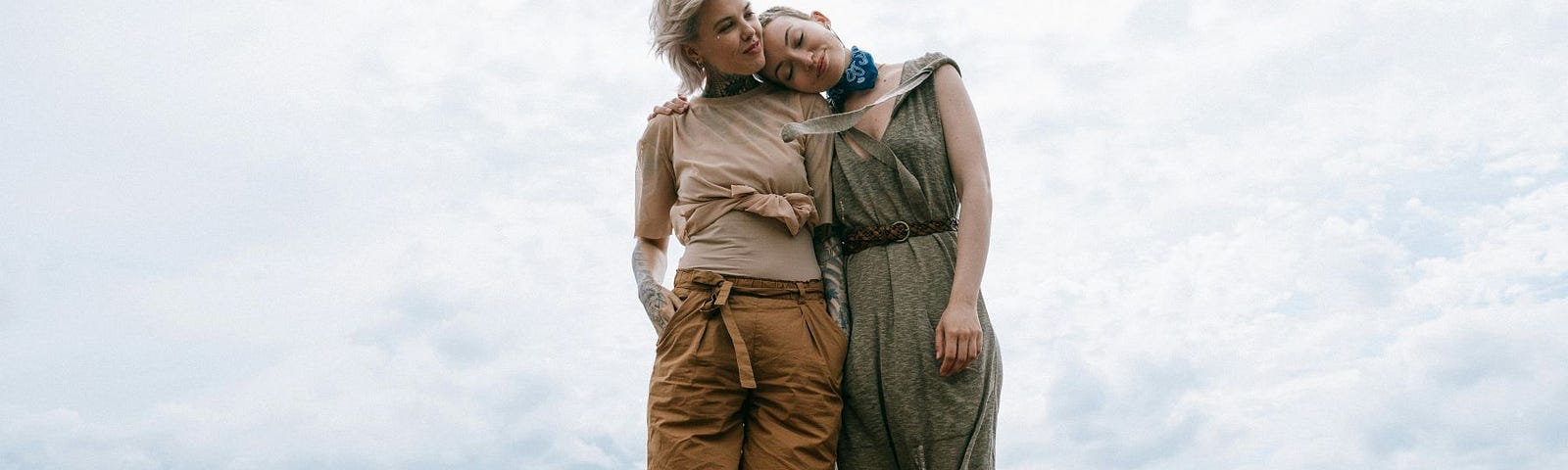 Two women standing on a dock with the water at their back, with one leaning her head on the other’s shoulder.