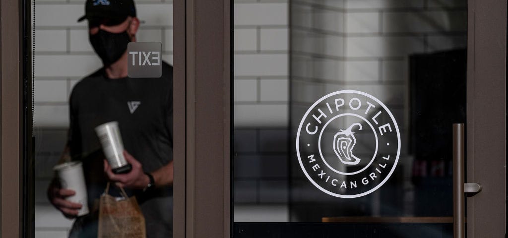 A masked employee behind a glass door with the “Chipotle Mexican Grill” logo