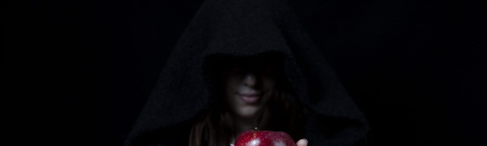 A woman cloaked in a hood is holding out an apple.