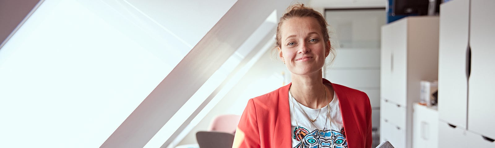 Tine Holm stands in the middle of a neutral work space, wearing a red blazer and holding a reMarkable paper tablet.