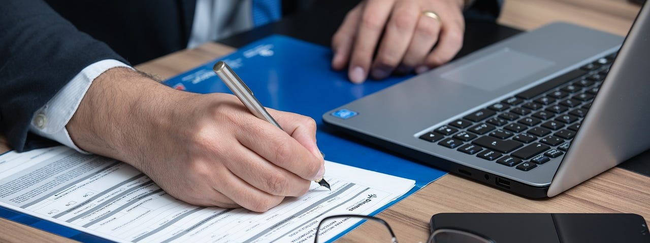 IMAGE: A person filling up details in a form while close to a laptop, with his glasses and a smartphone also on the table