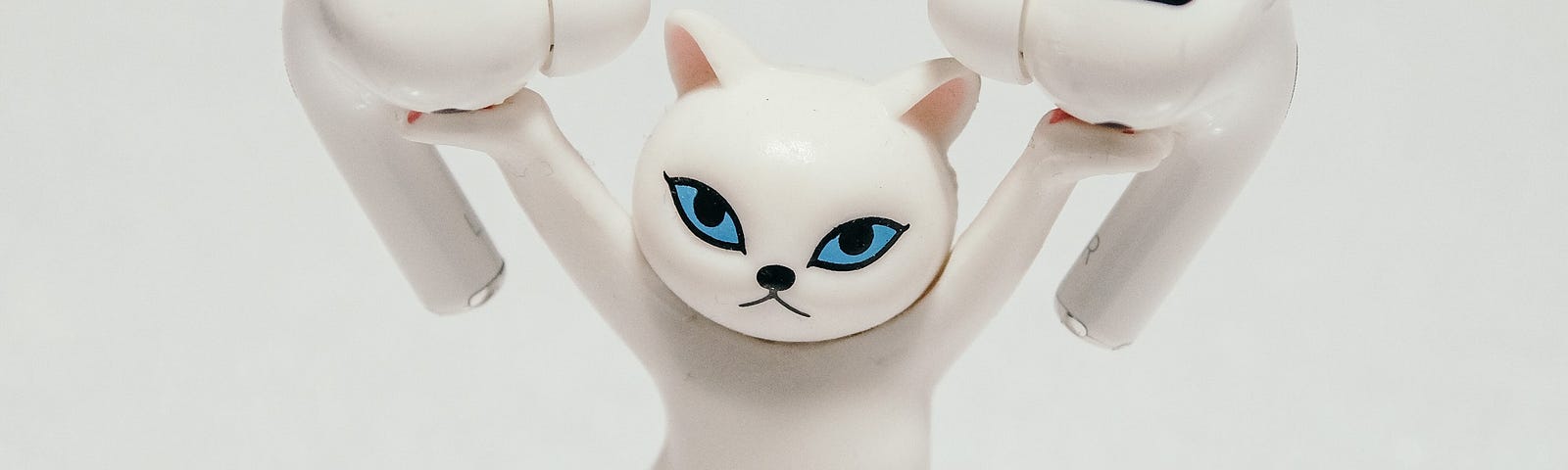 A white plastic cat with blue eyes is standing with its arms up and its right hip to the side. It holds a white wireless earbud in each of its hands. The cat is set on a white surface and its shadow can be seen directly behind its feet.