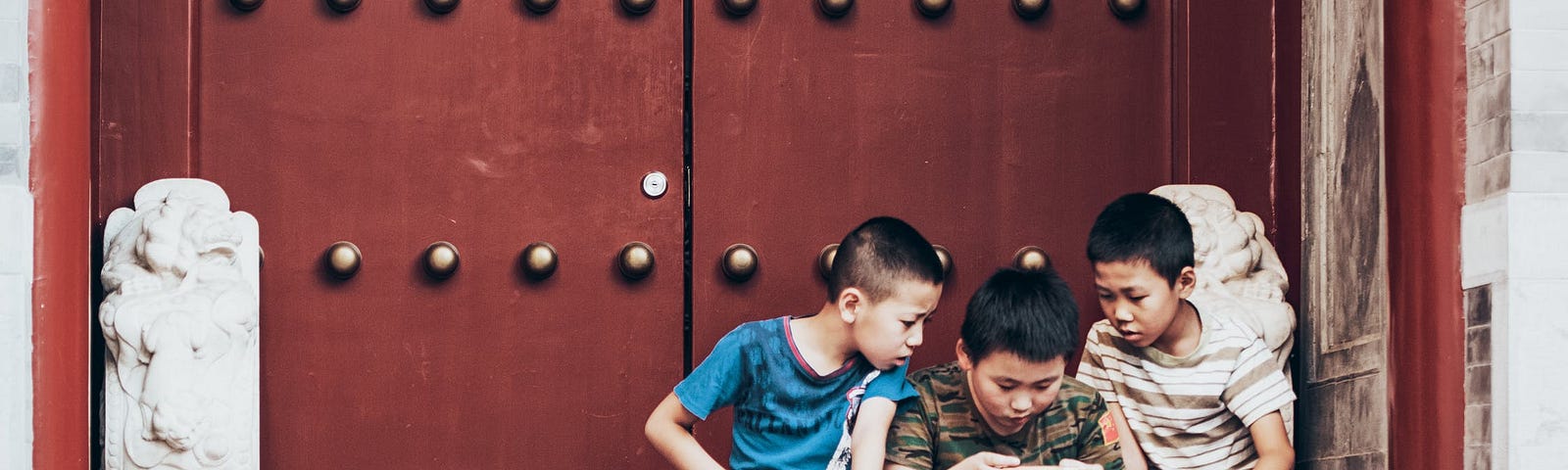 Three young Chinese boys sitting at the steps of a temple or ancient palace huddled over a single smartphone, with the two boys at the sides watching the one in the middle play a game.