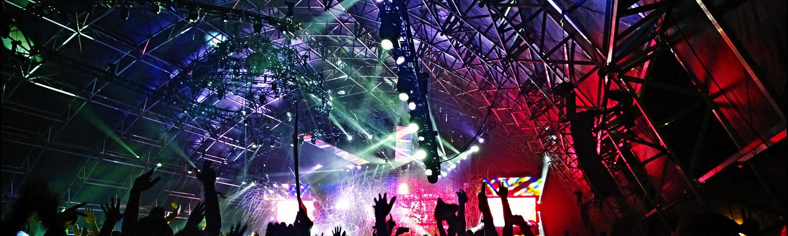 Silhoutted bodies at concert foregrounding colourful light-show on stage and semi-circle roof.