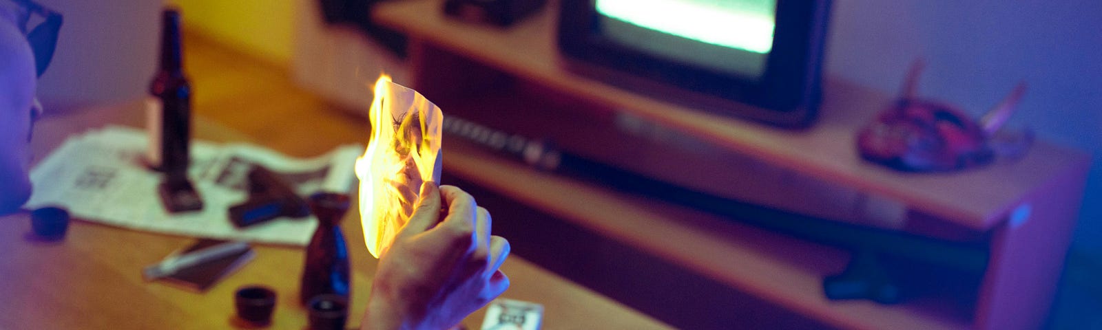 male burning a paper in a living room with a television in the background, a bottle on the table with an ashtray filled with cigarretts