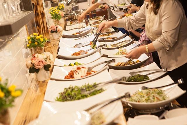 About 20 white buffet plates with different foodstuffs arrayed. People are serving themselves.