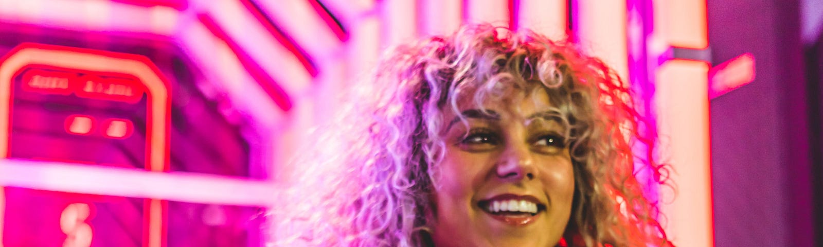 Curly-haired person in a hoodie smiling, lit up by pink arcade lights