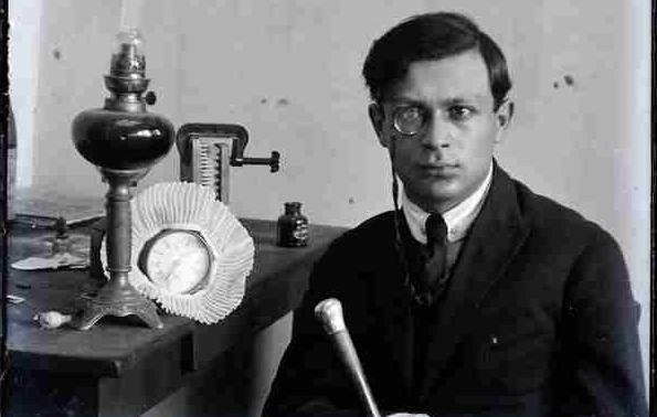 A black and white photo from the early 20th century of a man with light skin and dark hair wearing a suit and a monocle. He sites in front of a lamp and art materials.