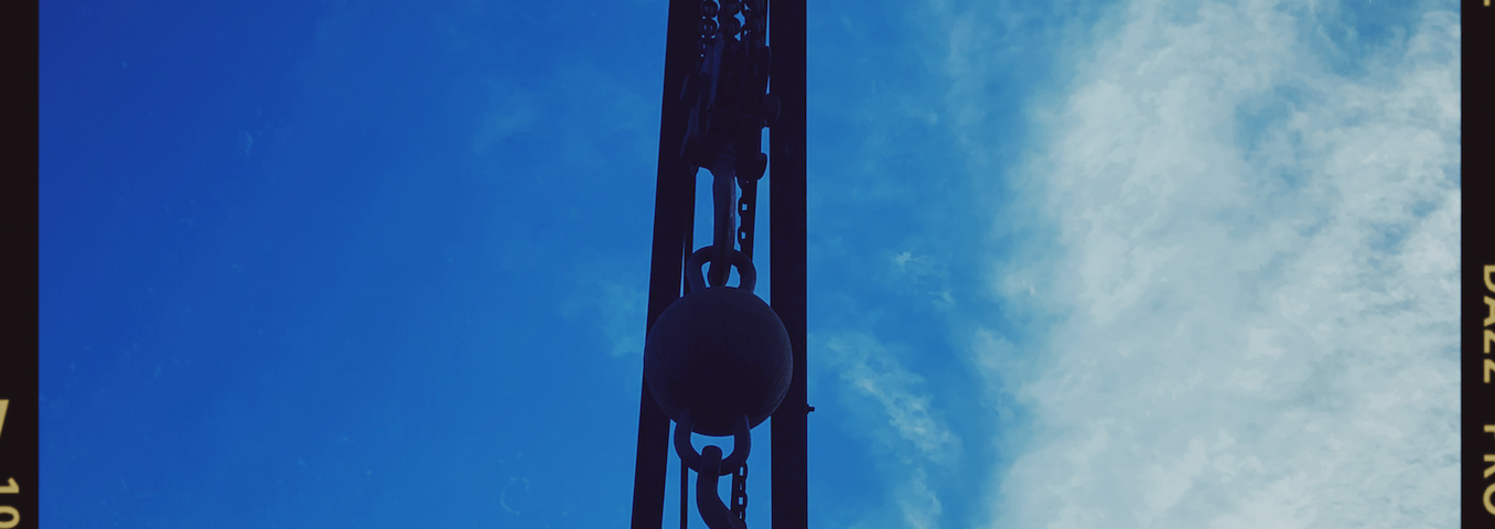 view from below a crane. it is stark black against a bright blue sky. There is a a hook with a ball hanging from it.