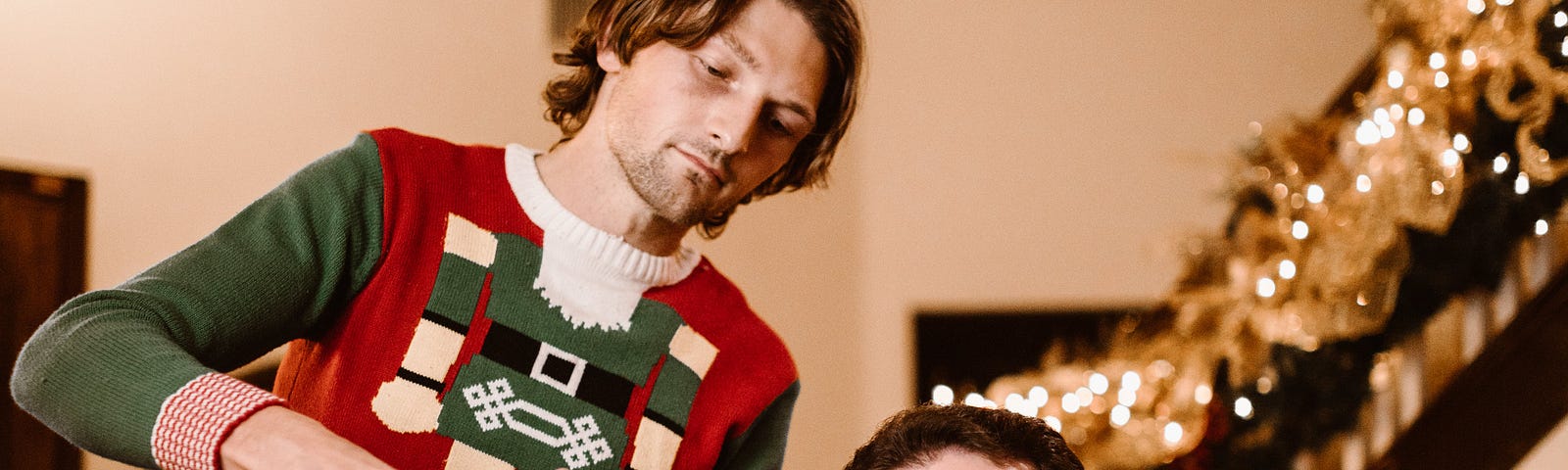 Two men in holiday sweaters, one pouring wine into the other’s glass while the one holding the glass makes a disgusted face.