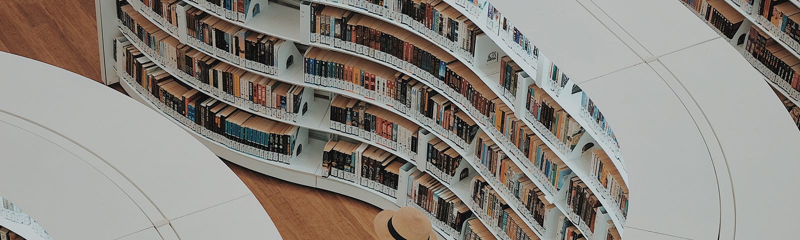 Photo taken from above of a woman in a hat looking at undulating, well stocked white bookshelves in a bookshop