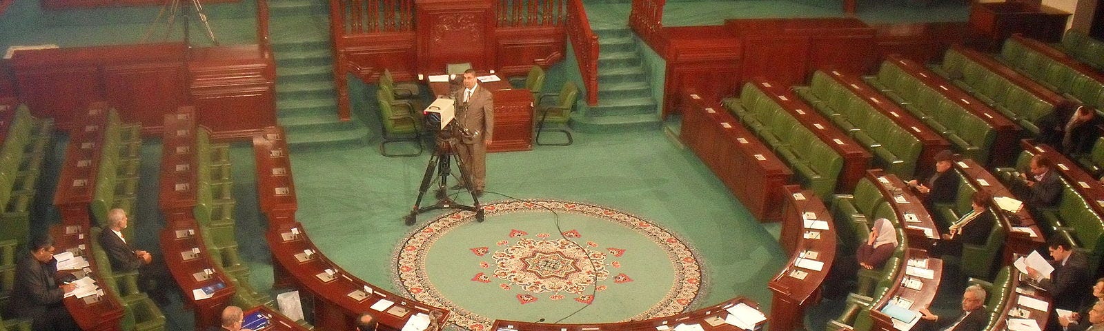 Tunisian Parliament in a horse shoe shape, with parliamentarians and a TV camera looking on.