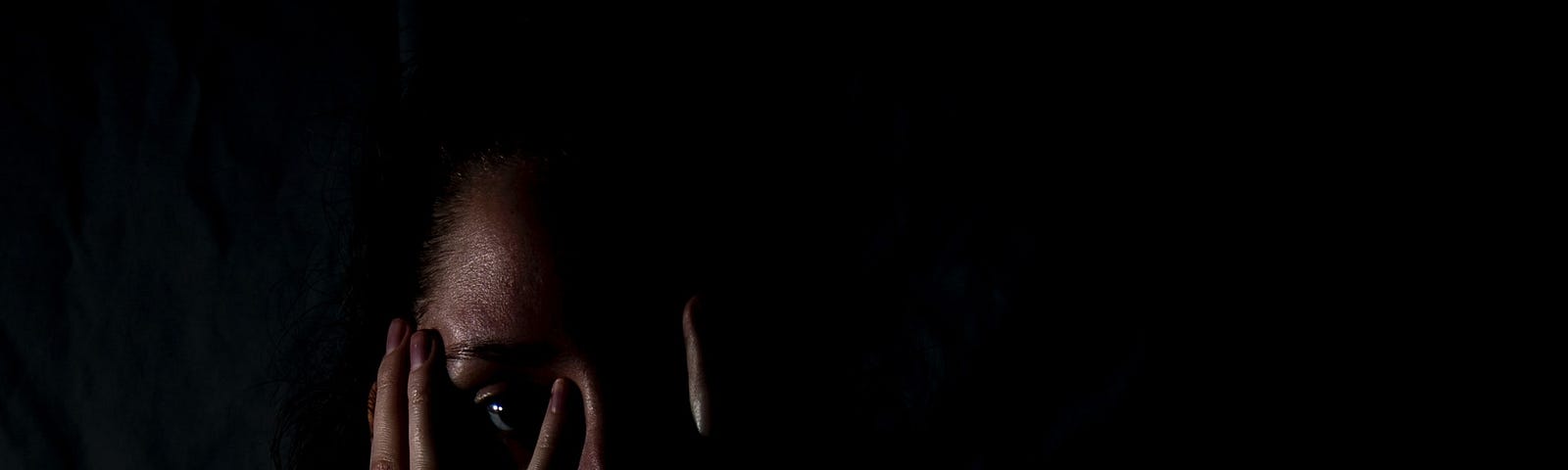 Against a black background, and in very low lighting, a woman covers her face with her hands, but spreads her fingers so she can see.