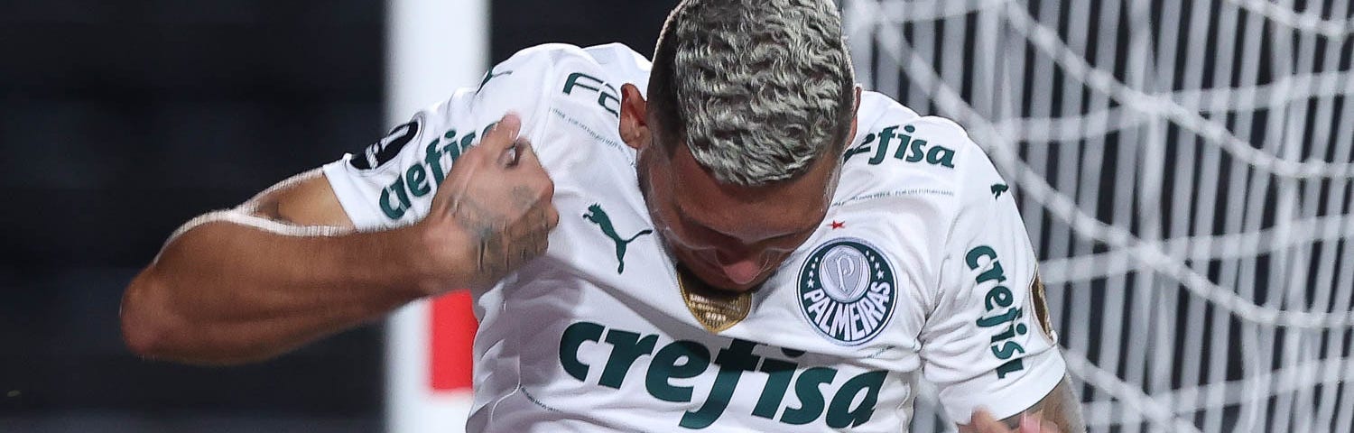 Rafael Navarro, atacante do Palmeiras, comemora gol em jogo pela Libertadores. Ele veste camisa branca, com detalhes em verde na horizontal, bem finos, e tem cabelo platinado. Ele faz movimento dos braços de cima para baixo, como quem “espanta a zica”