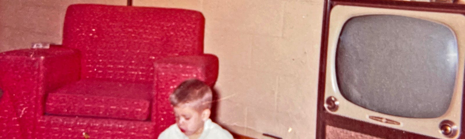 Boy sits on the floor playing with a toy train set. He’s sitting next to a large old style television and in front of a red tufted chair.