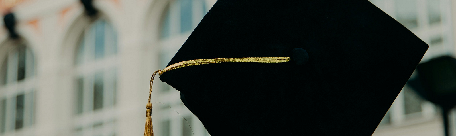 A hand holds a graduation cap in the air, in front of some lovely, old looking building.