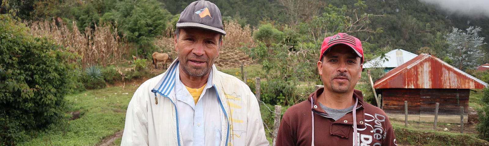 An image of two men in a rural location in Guatemala. They are standing in the foreground. In the background are some buildings in cleared areas among the vegetation. In the far background are hills partially shrouded in mist.
