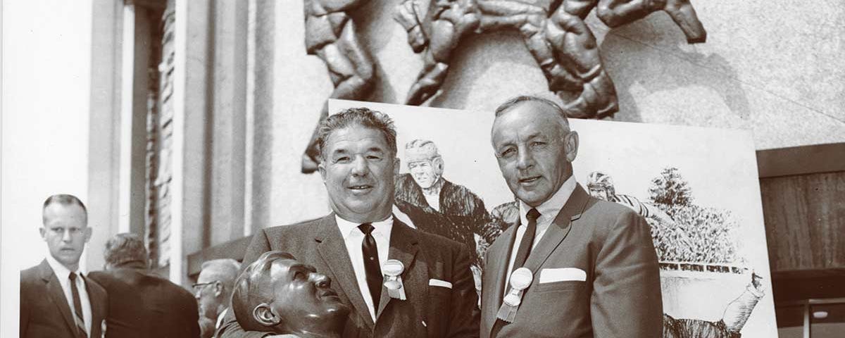 black and white photo of Arnie Huber holding his own bust and posing for a photographi with a man in a suit during Huber’s Hall of Fame induction ceremony.