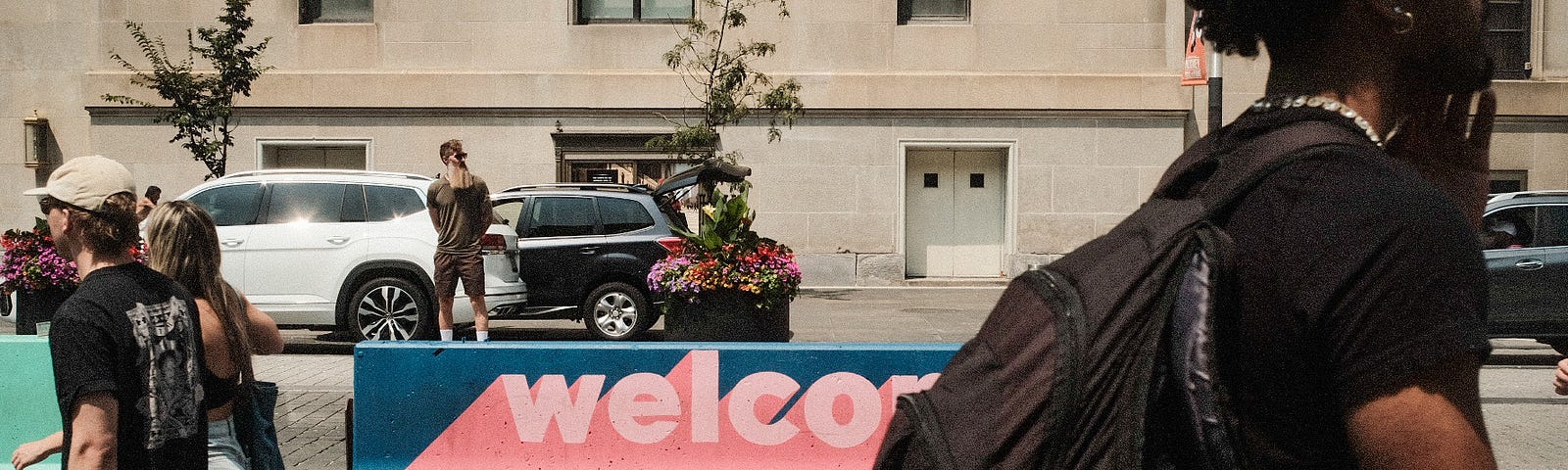 Pedestrians moving along the street, with a colored concrete barrier in the center of the frame that reads, “welcome.”