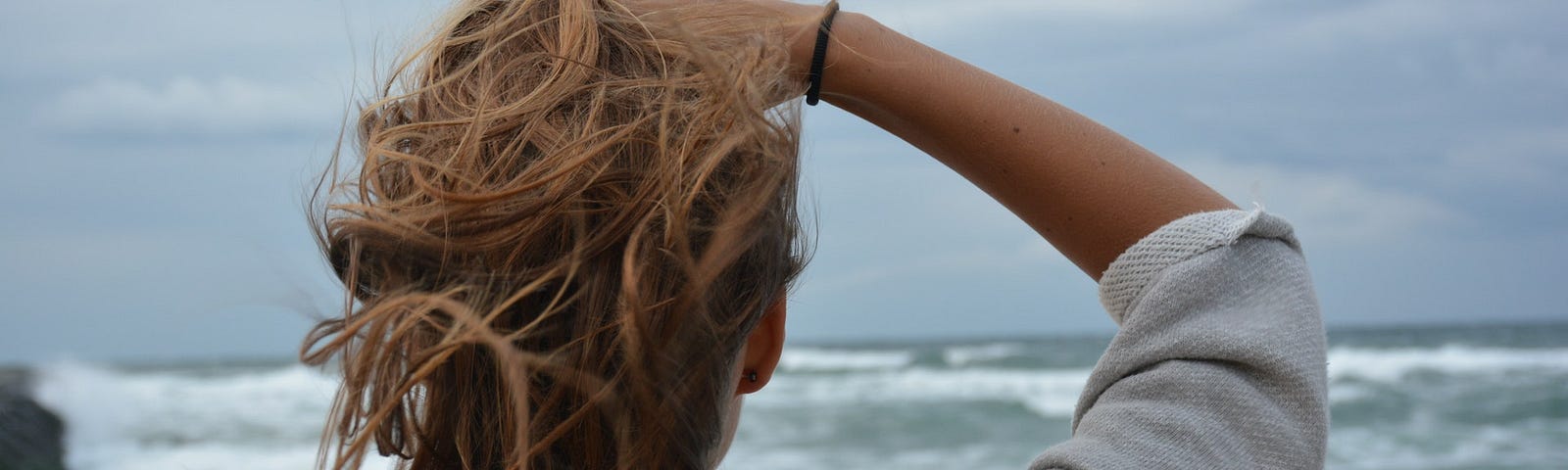 Back of woman with reddish-brown hair facing a cloud sky and turbulent ocean.