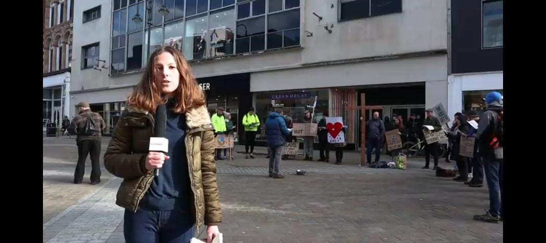 The author, a young woman, dressed in navy with a kaki jacket on, reporting in front of a crowd of protesters, holding a big microphone.