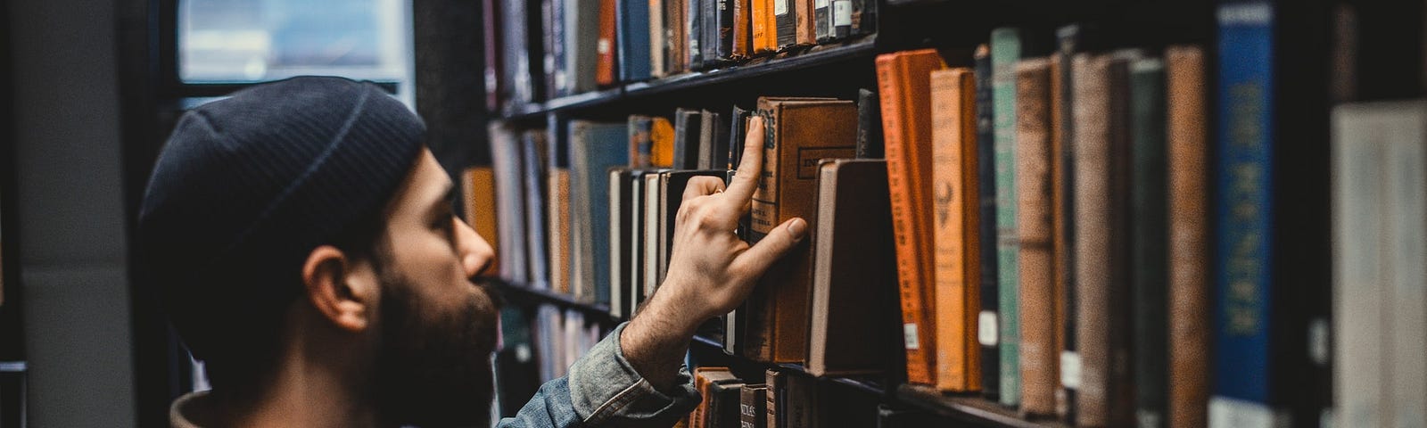 Nesta imagem temos um homem de touca na cabeça de frente a uma estante de biblioteca, estante alta onde tem vários livros, ele está com a mão levantada pegando um livro nessa estante.