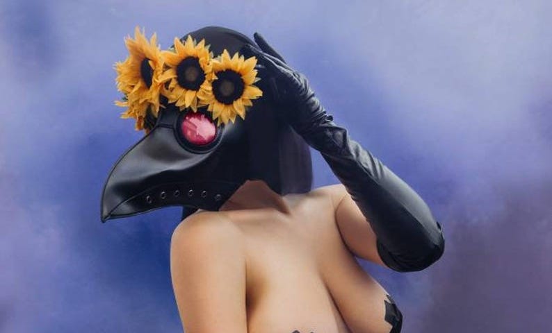 White female figure stands amidst clouds of grey and muted blue and purple, She wears a black leather plague doctor mask, her face completely obscured, with a bright yellow crown of sunflowers; a stark contrast to the bleak background, complimenting her bare skin. One leather gloved hand touches gently to the temple of the mask, the other falling to her bare thigh clad in leather and chain detailing.