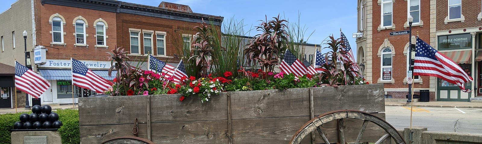 This shows an old wagon with American flags on a green lawn in front of old buildings.