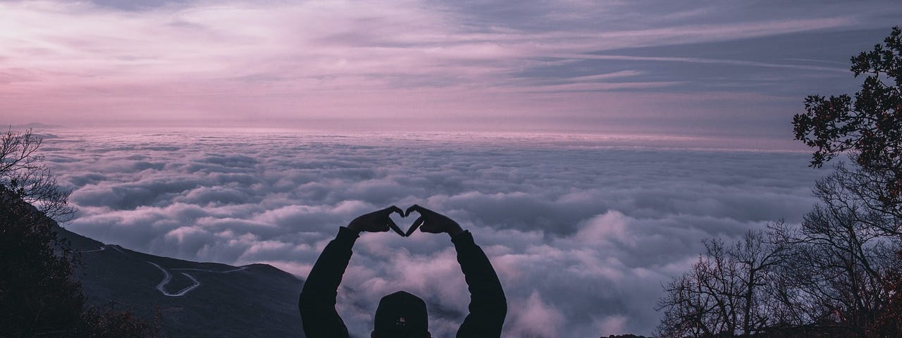 Person with hands in heart shape, overlooking low-level clouds from higher elevation.