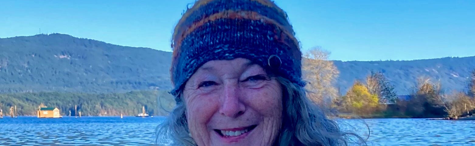 An older woman with a wool sweater and hat sits looking into the camera. The sun is shining and the sky is bright. She is smiling. Ocean water ripples behind her.