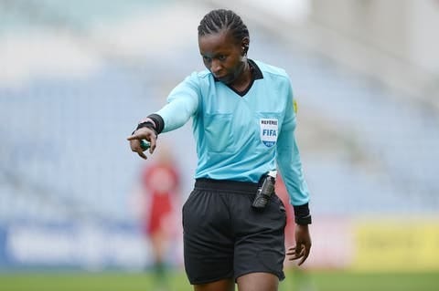 mulher negra escura de trança nagô com blusa azul clara com logo da FIFA e bermuda preta. ela está apontando para frente.