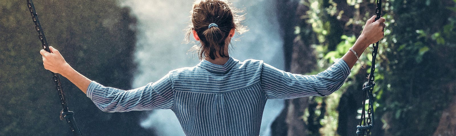 Back view of woman wearing long sleeve striped shirt sitting on swing overlooking canyon and rushing river