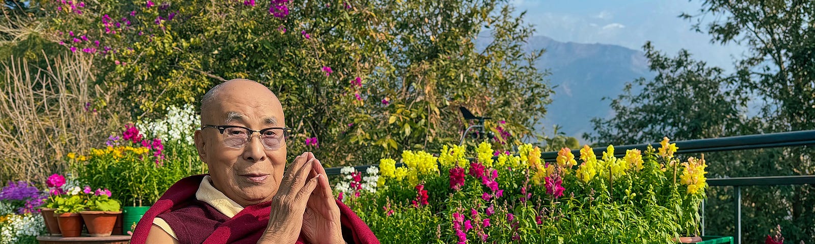 The Dalai Lama sitting on his patio in Dharamsala, India with colorful flowers behind him.