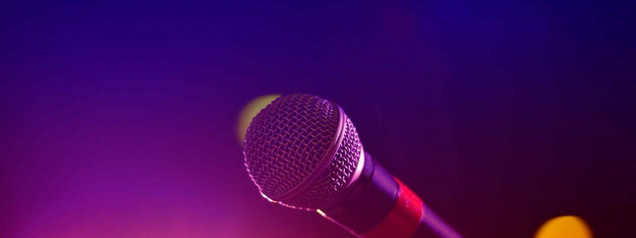 A microphone sits in a mic stand in the foreground. The background is purplish blue with some pink and yellow lights at the bottom.