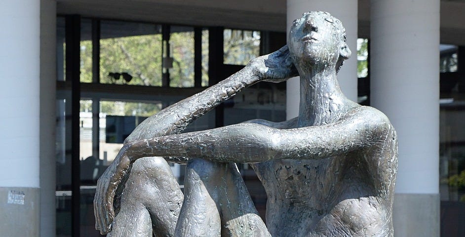 Statue of a naked man seated on the ground with his feet flat on the ground near the torso. The man is looking up with his left arm resting across his knees. The right elbow is on the right knee, the right hand is cupping the right ear.