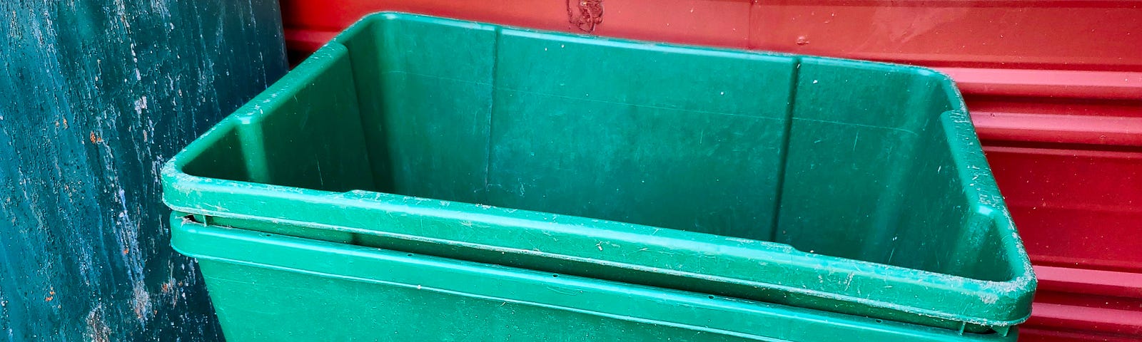 Two green recycling bins stacked on inside the other are placed against a red exterior wall and a green door. The bottom bin appears worn with a message encouraging recycling and an emblem representing sustainability.