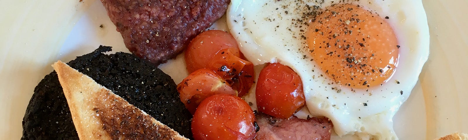 Breakfast, with fried egg, bacon, square slice sausage, triangular slices of toast, black pudding, tomatoes