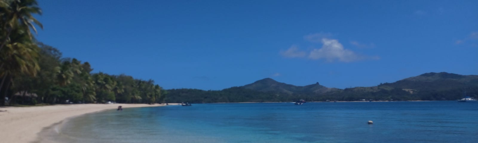 A tropical island beach with blue waters, white sand, boats near shore.